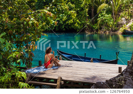 Woman in swimsuit sitting on a wooden pier by a turquoise lagoon, surrounded by lush tropical nature. 126764950
