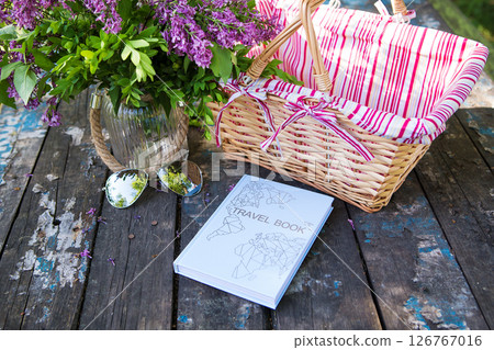 An idyllic still life featuring a travel book, sunglasses, and a vase of lilac flowers, set on an old wooden table next to a picnic basket 126767016