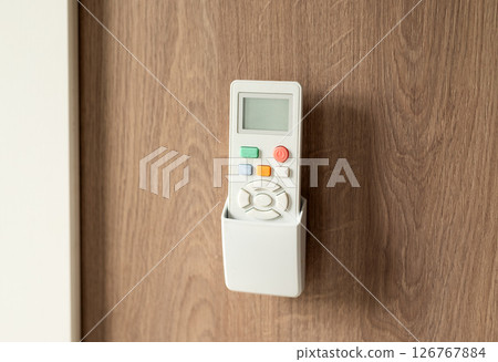 White remote control in a wall holder mounted on a wooden surface, used for air conditioning or home devices. 126767884