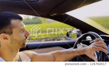 Profile of young handsome man driving a modern electric car with sunlight at background. Young male driver riding on electrical vehicle at countryside. Concept of journey and trip. Close up 126768017