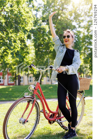 Portrait young adult pretty woman red vintage retro bike and pin-up casual outfit waving hand green city park bright sunny summer day. Happy beautiful female person cycling outdoor. Healthy lifestyle Portrait young adult pretty woman red vintage retro bike and pin-up casual outfit waving hand green city park bright sunny summer day. Happy beautiful female person cycling outdoor. Healthy lifestyle 126769294
