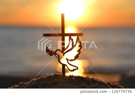 Black silhouette of stick in shape of cross and dove with branch with leaves in beak on background of sea, sea waves, sunny path and shining sun during sunrise or sunset with rising setting sun Black silhouette of stick in shape of cross and dove with branch with leaves in beak on background of sea, sea waves, sunny path and shining sun during sunrise or sunset with rising setting sun 126770736