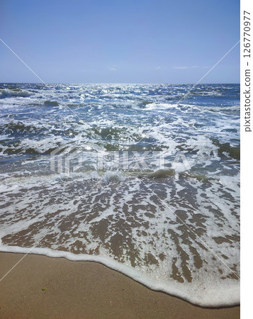 Clean transparent waves of sea with white foam and sun glare rolling onto sandy beach of seashore on sunny summer day with blue sky. Seashore coast. Sandy beach. Nature Natural background Clean transparent waves of sea with white foam and sun glare rolling onto sandy beach of seashore on sunny summer day with blue sky. Seashore coast. Sandy beach. Nature Natural background 126770977