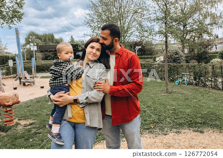 Young family spending time together outdoors in a park. Mother holds a toddler while father embraces them, all dressed in casual clothes, enjoying a peaceful day in nature. 126772054