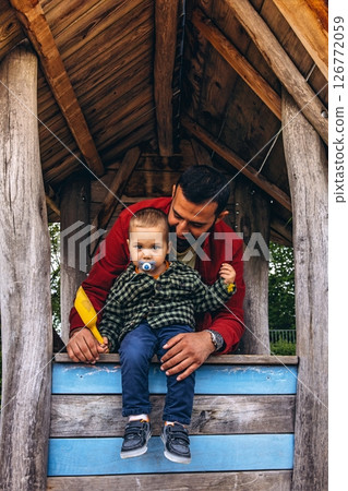 Father and young child playing together in a wooden playhouse at an outdoor playground. The child pours sand from a yellow scoop, surrounded by natural wood textures and greenery 126772059
