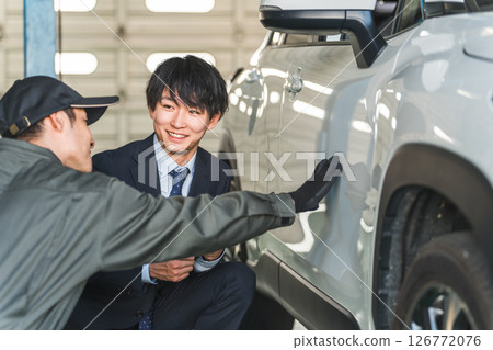 A male mechanic and a salesman in a suit checking scratches and dents on a car A male mechanic and a salesman in a suit checking scratches and dents on a car 126772076