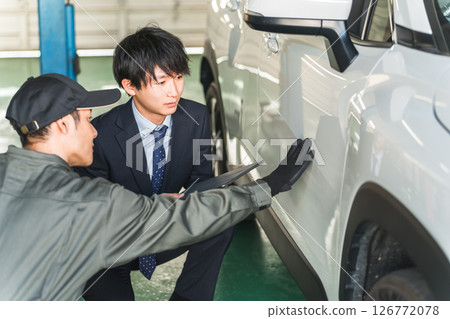 A male mechanic and a salesman in a suit checking scratches and dents on a car A male mechanic and a salesman in a suit checking scratches and dents on a car 126772078