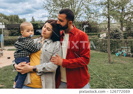 Young family spending time together outdoors in a park. Mother holds a toddler while father embraces them, all dressed in casual clothes, enjoying a peaceful day in nature. Young family spending time together outdoors in a park. Mother holds a toddler while father embraces them, all dressed in casual clothes, enjoying a peaceful day in nature. 126772089