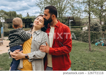 Young family spending time together outdoors in a park. Mother holds a toddler while father embraces them, all dressed in casual clothes, enjoying a peaceful day in nature. 126772118