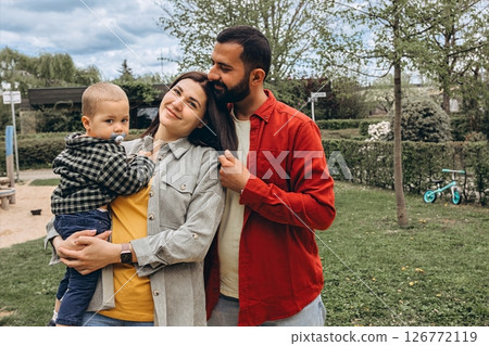 Young family spending time together outdoors in a park. Mother holds a toddler while father embraces them, all dressed in casual clothes, enjoying a peaceful day in nature. 126772119