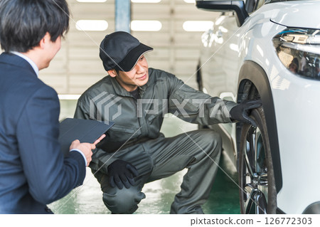 A male car mechanic and a front man/salesman in a suit having a conversation in front of a car 126772303