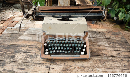 Rusty Vintage Typewriter with Dusty Keys and Weathered White Casing Sitting on an Aged Wooden Table in an Outdoor Setting with Brick Pathway and Green Plants in the Background Rusty Vintage Typewriter with Dusty Keys and Weathered White Casing Sitting on an Aged Wooden Table in an Outdoor Setting with Brick Pathway and Green Plants in the Background 126776656