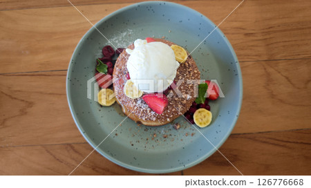 Fluffy pancakes with vanilla ice cream, strawberries, bananas, red berries, mint, and crushed nuts on a pastel green plate over a wooden table. 126776668