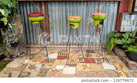 Three Handmade Industrial Bar Stools Repurposed from Half Gas Cylinders with Metal Legs in a Rustic Outdoor Setting Featuring Corrugated Metal Wall and Colorful Mosaic Tile Flooring with Green Plants Three Handmade Industrial Bar Stools Repurposed from Half Gas Cylinders with Metal Legs in a Rustic Outdoor Setting Featuring Corrugated Metal Wall and Colorful Mosaic Tile Flooring with Green Plants 126776672