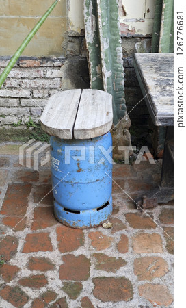 Outdoor handmade stool creatively repurposed from a blue gas cylinder with a wooden seat on a rustic brick patio near weathered walls and green cactus plants. 126776681