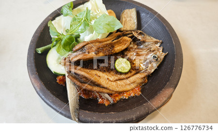 Crispy fried milkfish served with spicy sambal penyet and fresh vegetables on a rustic black plate in Indonesia featuring vibrant garnishes and traditional presentation. 126776734
