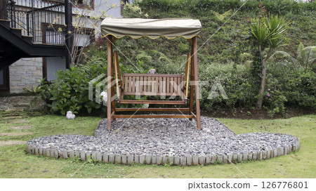 Wooden garden swing with a beige canopy placed on a pebble platform surrounded by lush green plants and a hillside backdrop in a peaceful outdoor setting Wooden garden swing with a beige canopy placed on a pebble platform surrounded by lush green plants and a hillside backdrop in a peaceful outdoor setting 126776801