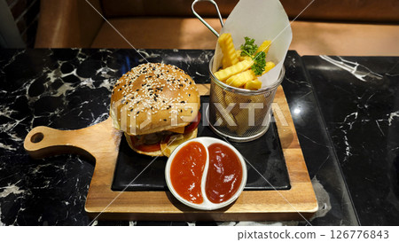 Juicy beef burger with sesame seed bun served on a wooden tray with crispy French fries and dipping sauces on a marble table in a cozy restaurant setting Juicy beef burger with sesame seed bun served on a wooden tray with crispy French fries and dipping sauces on a marble table in a cozy restaurant setting 126776843