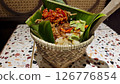 Traditional Nasi Bakul with red chili sambal, fried shallots, and petai served in a woven bamboo basket lined with banana leaves on a colorful terrazzo table in an Indonesian dining setting 126776854