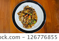 Stir-fried flat rice noodles with rich soy sauce, fresh vegetables, and red chilies served on a white plate with a black rim on a wooden table in a vibrant setting. 126776855