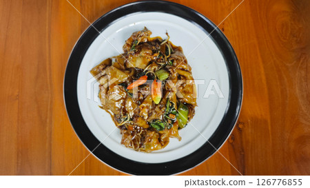 Stir-fried flat rice noodles with rich soy sauce, fresh vegetables, and red chilies served on a white plate with a black rim on a wooden table in a vibrant setting. 126776855