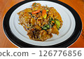 Stir-fried flat rice noodles with rich soy sauce, fresh vegetables, and red chilies served on a white plate with a black rim on a wooden table in a vibrant setting. 126776856