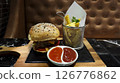 Juicy beef burger with sesame seed bun served on a wooden tray with crispy French fries and dipping sauces on a marble table in a cozy restaurant setting 126776862