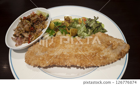 Crispy breaded fish fillet served with sauteed chili onions roasted potatoes fresh green salad and parsley garnish on a white plate creating a vibrant and appetizing dish presentation. 126776874