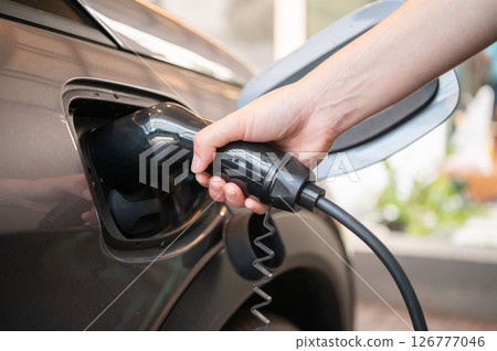 Cropped shot view of EV car driver while inserts the electrical connector to charge the batteries of EV car. Cropped shot view of EV car driver while inserts the electrical connector to charge the batteries of EV car. 126777046