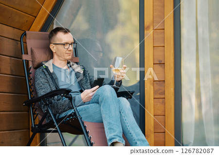 Man looking at smartphone sitting on terrace of a house with a glass of wine 126782087