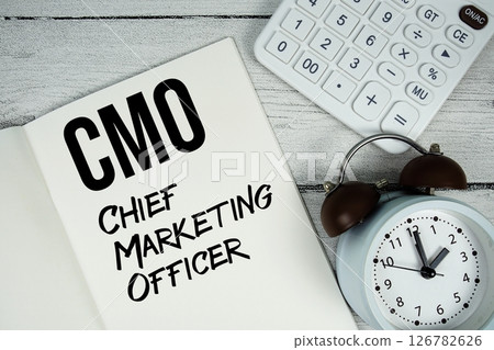 CMO Chief Marketing Officer text on paper notebook page with alarm clock and calculator on wooden background CMO Chief Marketing Officer text on paper notebook page with alarm clock and calculator on wooden background 126782626