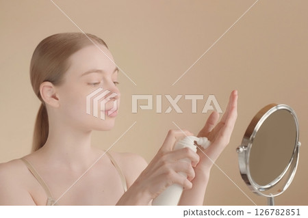 Face. Young woman applying skincare foam on her hand in front of small mirror on isolated light background. Beautiful model girl. Perfect clear skin. Selfcare concept, beauty cosmetics advertisement Face. Young woman applying skincare foam on her hand in front of small mirror on isolated light background. Beautiful model girl. Perfect clear skin. Selfcare concept, beauty cosmetics advertisement 126782851