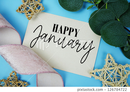 Happy January text on paper card with fabric ribbon and rattan star decorations on blue background Happy January text on paper card with fabric ribbon and rattan star decorations on blue background 126784722