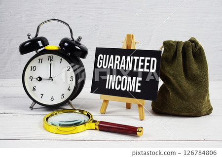 Guaranteed Income text with alarm clock and magnifying glass on wooden background Guaranteed Income text with alarm clock and magnifying glass on wooden background 126784806