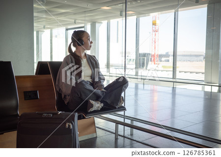 A woman sits cross-legged on an airport bench, listening to music through headphones and looking out the window, suitcase nearby. Concept of peaceful travel, anticipation and solo journey experience. 126785361