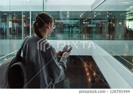 Young woman with a backpack holding a passport and smartphone while looking through glass at a contemporary airport. Concept of international travel, identity verification and global mobility. 126785494