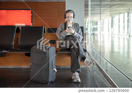 A young woman sits on a bench in a bright airport terminal, wearing headphones and looking at her smartphone with suitcase nearby. Waiting during international travel and digital technology. 126785600