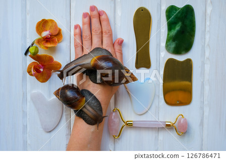 Two achatinas on hand and facial massage kit. Face roller and guasha massager made from natural stones and metal, flat lay. High quality photo Two achatinas on hand and facial massage kit. Face roller and guasha massager made from natural stones and metal, flat lay. High quality photo 126786471