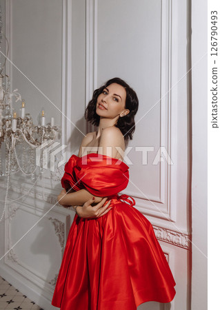 Woman in red off-shoulder dress posing by chandelier in classic white room 126790493