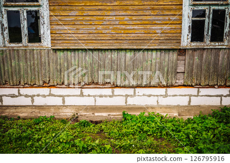 Old wooden wall with damaged planks and two windows showing foundation and grass Old wooden wall with damaged planks and two windows showing foundation and grass 126795916