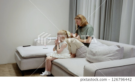 Caring mother gently brushing daughter's hair, sharing tender bonding moment on living room sofa, expressing maternal love and connection Caring mother gently brushing daughter's hair, sharing tender bonding moment on living room sofa, expressing maternal love and connection 126796442