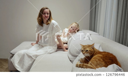 Loving mom and small daughter relaxing on comfortable sofa, cuddling ginger cat, sharing quiet family moment with tender connection and domestic warmth 126796446