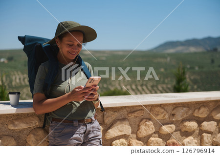 Traveler in Sun Hat and Backpack Enjoying Mobile Phone Among Scenic Landscape Traveler in Sun Hat and Backpack Enjoying Mobile Phone Among Scenic Landscape 126796914