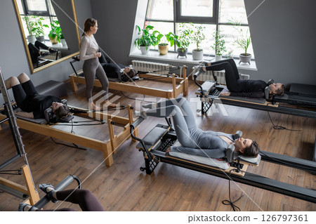 Group of caucasian women exercising with trainer on pilates reformers in a bright studio. Their workout focuses on core strength and flexibility in a modern fitness environment 126797361