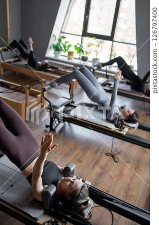 Group of caucasian women exercising with trainer on pilates reformers in a bright studio. Their workout focuses on core strength and flexibility in a modern fitness environment 126797400