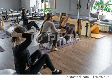 Group of women doing exercises on back spine corrector. Reformer pilates studio machine for fitness workouts in gym. Fit, healthy and strong authentical body. Fitness concept. 126797426