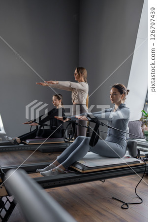 Group of caucasian women exercising with trainer on pilates reformers in a bright studio. Their workout focuses on core strength and flexibility in a modern fitness environment 126797439