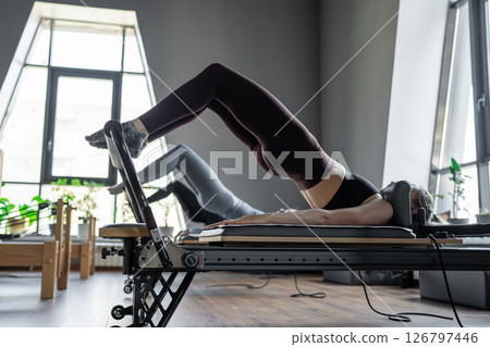 A group of woman exercises Glute bridge on a reformer bed machine in natural sunlight, focusing on pelvis and hips. 126797446