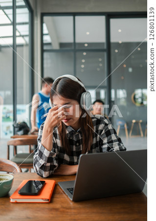 Woman in headphones looking stressed at a laptop in a modern office setting, with a smartphone and notebook nearby. 126801139