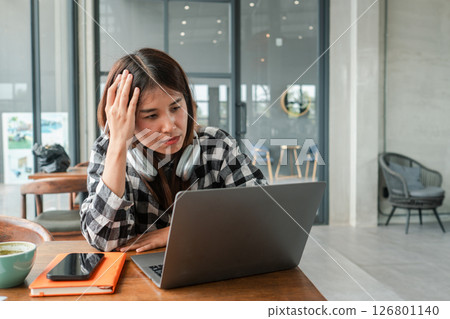 Focused woman using laptop in a stylish cafe, wearing headphones, with a smartphone and notebook nearby. 126801140